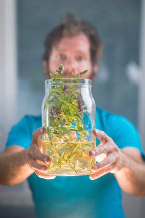 Different herbs or green plants in a glass transparent jar with some water in it, held by a man in...