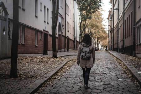 Lonely woman with a backpack walking away from camera on a lonely path or alley in a city. Dangerous feeling or solitude in the city.の写真素材