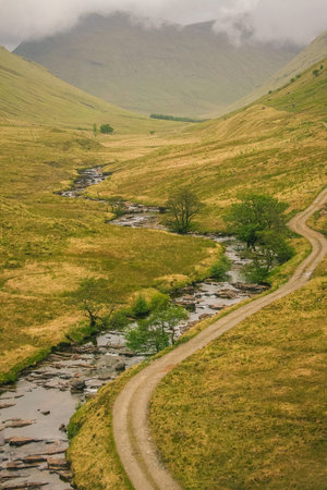 Vertical panorama of a typical scottish nature, with a fire road and a small river on a lush green...