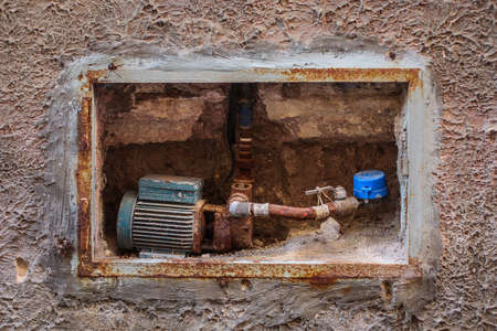 An old electric motor water pump installed in a wall of a house. Old rusty vintage water pump with visible piping, valve and counter. Concept of old piping systems in houses.の写真素材