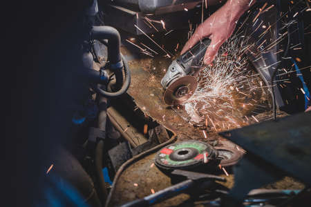 Bare hands of a man using an angle grinder to cut off the excess rusty metal from a car body in the attempt to restore it and weld in a new piece. Sparks are flying around, tools and grinding plates lying around.の写真素材