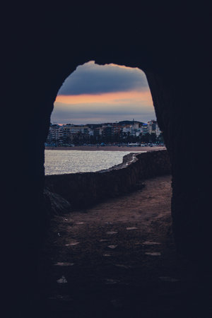 Looking through a rock tunnel on a pedestrian walkway towards sandy beachfront on popular tourist destination Lloret De Mar in Spain in evening with sun slowly falling behind the buildings.の写真素材