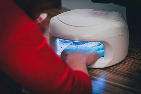 Hand in the UV dryer, to dry and harden gel varnish for nails. Woman in red shirt is having her...