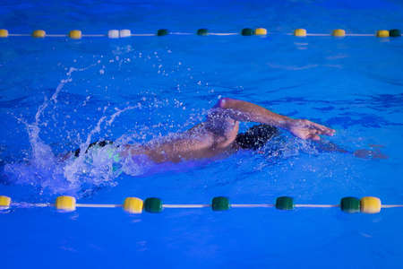 A person swimming freestyle stroke in an indoor pool lit with colorful underwater lights. Visible hand and arm of a swimmer, surrounded by drop and droplets.の写真素材