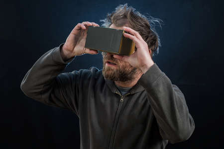 A hipster looking man looking through a cardboard VR glasses with a curious expression on his face on a dark black lit background.の写真素材
