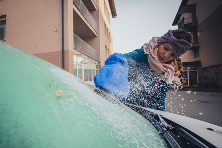 Woman is cleaning an icy window on a car with ice scraper designed as a glove. Ice and snow chunks...