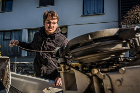 A young male is trying to start an outboard engine with pulling the rope on the starter. Industrial houses in the background.の写真素材