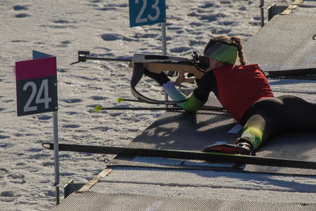 Female biathlon racer is lying on the ground and aiming her rifle. Biathlete woman on a shooting range, firing lying down.の写真素材
