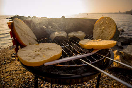 Grilling burgers on a small metal portable charcoal grill on the shores of Lac Leman in Switzerland during early winter evening with sun setting down. Visible cheese, meat and buns.の写真素材