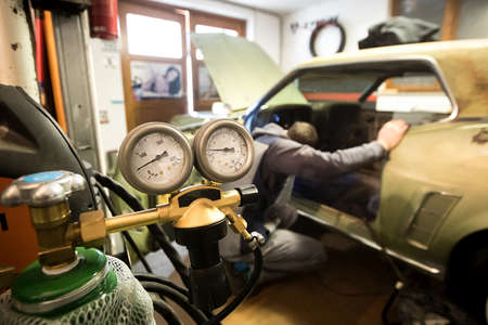 Welding tank pressure gauges are seen in the foreground while a  man is inspecting an old vintage car with an attempt to restore it.の写真素材