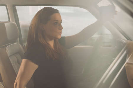 Young caucasian woman is happy driving a vintage sport car and adjusting the rear view mirror. Side view in a car, fast blurred image of a city in the background.の写真素材