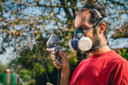 Amateur painter in red sweater and face mask using a spray gun to paint a plastic object or part in a home garden. Profile view of a painter with a small spray gun.の写真素材