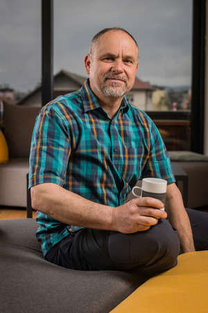 A caucasian man in his late forties with short hair and short gray beard is sitting in a couch and looking into the camera while holding a beverage cup. Forest is seen behind the window.の写真素材