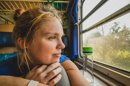 Caucasian woman using looking through a window of an older train with vintage seats and wooden walls. Woman commuting with an older retro train. Sun flare visibleの写真素材