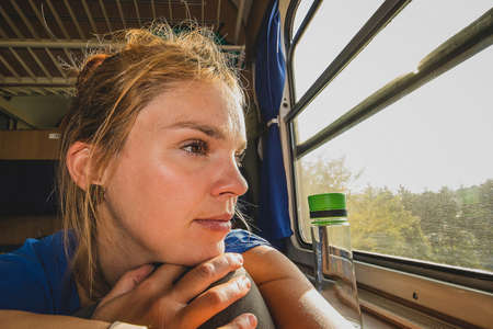 Caucasian woman using looking through a window of an older train with vintage seats and wooden walls. Woman commuting with an older retro trainの写真素材