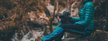 Side view of an unknown woman sitting on a rock outdoors in hiking clothes holding a photo camera. Waterfall and green nature in the background.の写真素材