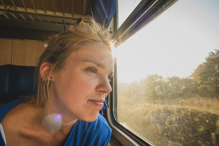 Caucasian woman using looking through a window of an older train with vintage seats and wooden walls. Woman commuting with an older retro train. Sun flare visibleの写真素材