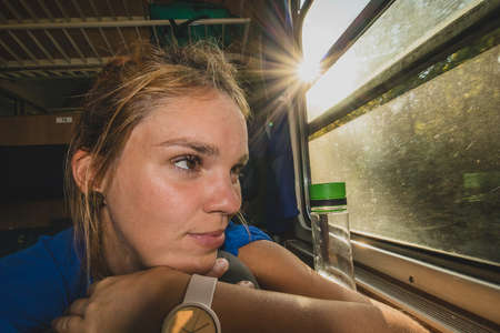 Caucasian woman using looking through a window of an older train with vintage seats and wooden walls. Woman commuting with an older retro train. Sun flare visibleの写真素材