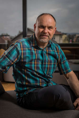 A caucasian man in his late forties with short hair and short gray beard is sitting in a couch and looking into the camera. Forest is seen behind the window.の写真素材