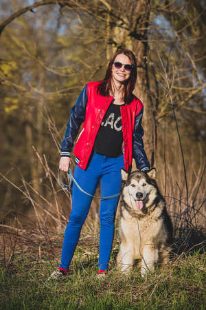 A girl with a smile  is standing with a siberian husky dog on a leash in a field with trees in early spring. Girl is wearing a red and blue leather jacket with blue pants.の写真素材