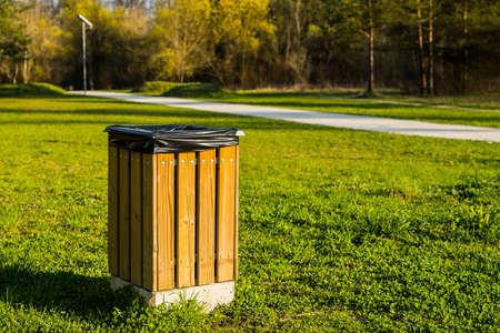 A modern wooden trash can or bin in a park with a black plastic bag visible inside on a sunny day. Walkway or path for pedestrians in the background.の写真素材