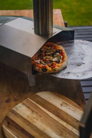 Man is pulling a delicious fresh home made pizza out of a stainless steel home portble oven fueled by pellets. Outdoor pizza party.の写真素材