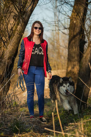 A girl with a smile  is walking with a siberian husky dog in a field and trees. Girl walking and a dog is rushing past her.の写真素材