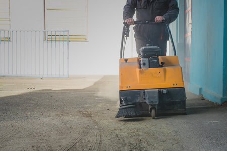 A person is pushing an orange gasoline powered industrial sweeper over dirty asphalt ground in an attempt to clean it from debris and sandの写真素材