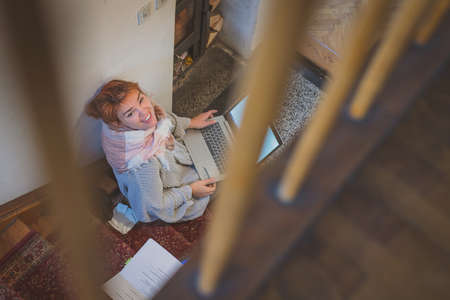 Young woman learning on the stairs next to a fireplace, holding a laptop and looking up. Romantic setting for learning in winter. Enjoying cozy winter learning session.の写真素材