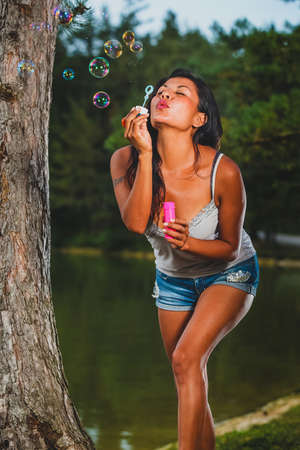 Playful young latin american woman is blowing party bubbles from soap outdoors with a lake in the background.の写真素材
