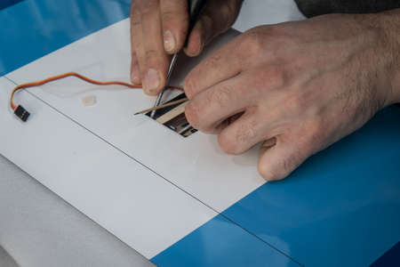 Hands of a person with tools assembling a servo RC motor in a wing of an aeroplane model. Wing in a blue color, connector wires seen in the background and wooden servo frame.の写真素材