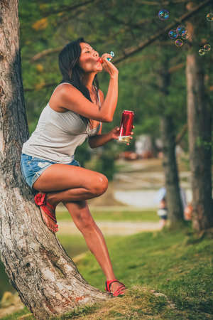 Playful young latin american woman is blowing party bubbles from soap outdoors with a lake in the background.の写真素材