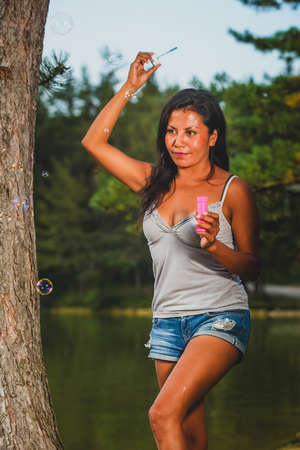 Playful young latin woman is blowing party bubbles from soap outdoors with a lake in the background.の写真素材