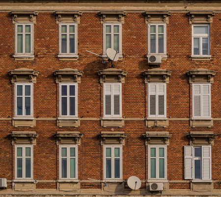 View of a brick facade with white windows. Two air conditioner units and satellite dishes on a sunny day.の写真素材