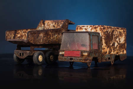 Pair of old, dirty and very rusty tin metal models of toy trucks in blue and grey color in a black and blue backgorund on reflective surface. One wheel is missing.の写真素材