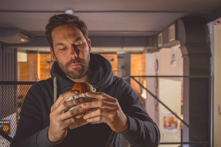 Young caucasian male with a beard holding a fresh hamburger and taking a big bite in a fast food...