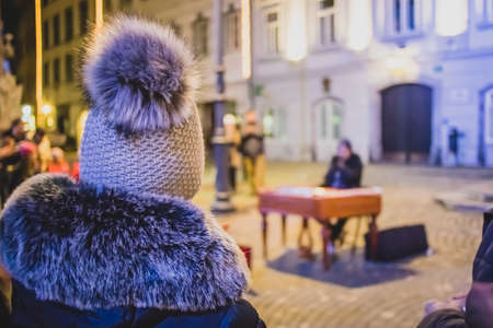 Back view of woman or a person with a white wool cap enjoying a public street performance in a city in winter. Street artist performing in front of a group of people.の写真素材