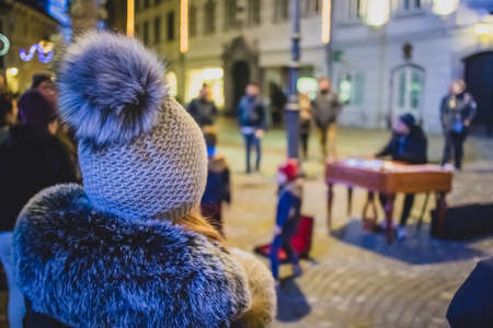 Woman enjoying a street performance at night while young kids are throwing coins in the bag of a street performer on a cold busy winter street.の写真素材