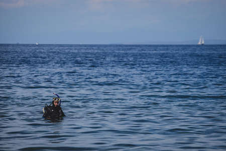A young female diver, in full gear to go underwater diving standing in the shallow water of a lagoon or sea with a mask and snorkel on her head.の写真素材