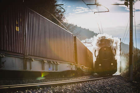 Old vintage steam train with black war time locomotive is running on a railroad track towards the camera on a sunny day. Another train is just passing by. Contrast of old and new.のeditorial素材