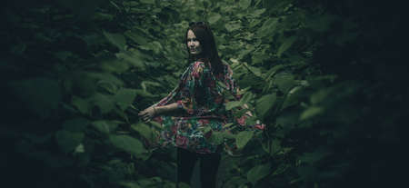 A young woman with red hair and sunglasses is posing and sitting down on a path full of leaves around her. Woman surrounded by lush greens and water reflection.の写真素材