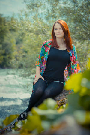 Cute redhead woman sitting next to a wild river, smiling closing her eyes and thinking about the future. Concept photo of a future dreamer.の写真素材