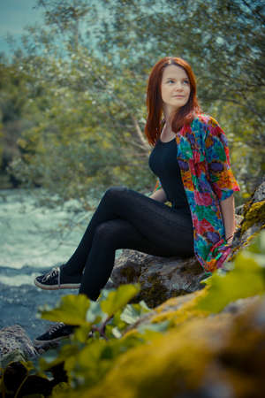 Portrait of a cute redhead woman  sitting next to a river surrounded by lush greens and looking into the distance thinking about the future.の写真素材