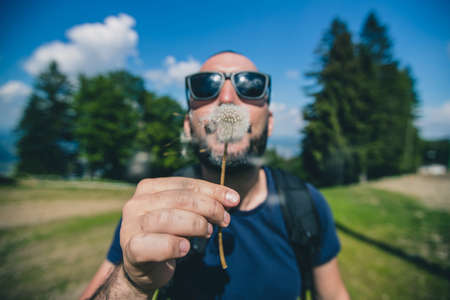 Frontal portrait of a handsome young bearded man blowing a dandelion flower in a high outdoor mountain alpine environment. Enjoying nature in spring while hikingの写真素材