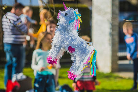 White colored donkey shaped pinata hanging on the tree with kids waiting to hit it in the background. Festive activity during a birthdayの写真素材