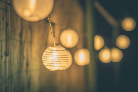 Row of round paper lanterns in a home garden on a wooden thatched background at night. Festive decoration in the garden on an evening.の写真素材