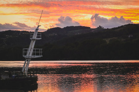 Picturesque and colorful sunset over a wide river of Drava close to Maribor, with the sun setting behind small green hills in the background. White mooring mast is seen on the shore.の写真素材