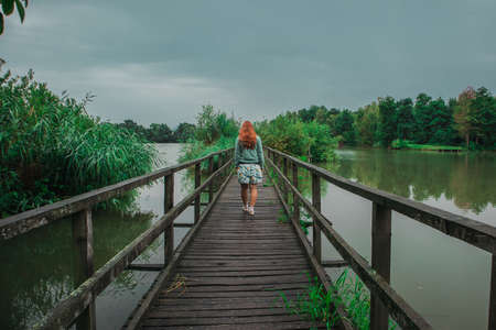 Back view of a female walking on a wooden bridge through green bushy lake with a lot of leaves on a murky scary cloudy day.の写真素材
