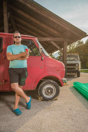 Man with a beer leaning on a side of a red vintage rusty and destroyed truck or van. Car restoration in progress. Tough guy restoring a car, drinking a beer. Another vintage van in the background.の写真素材