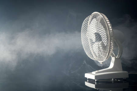 Side view of a white electric desktop fan on a black background and reflective floor with visible fog or mist blowing through. Concept of freshness with a fanの写真素材